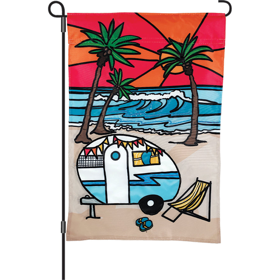 12 in. Beach Camper garden flag featuring a camper on the beach with waves and palm trees, displayed outdoors in a sunny garden setting.