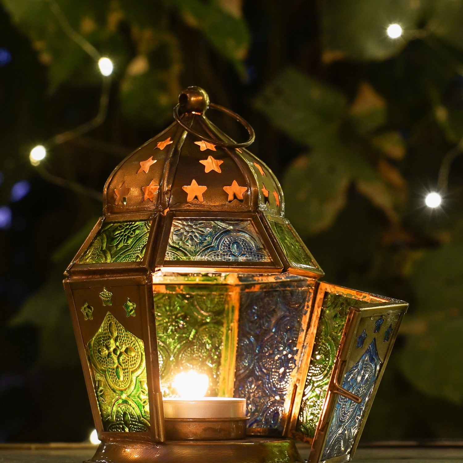 Blue & Green Hex Glass Tealight Lantern hanging in a garden, showcasing textured panels and an antiqued brass top, designed for tealight candles.