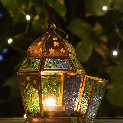 Blue & Green Hex Glass Tealight Lantern hanging in a garden, showcasing textured panels and an antiqued brass top, designed for tealight candles.
