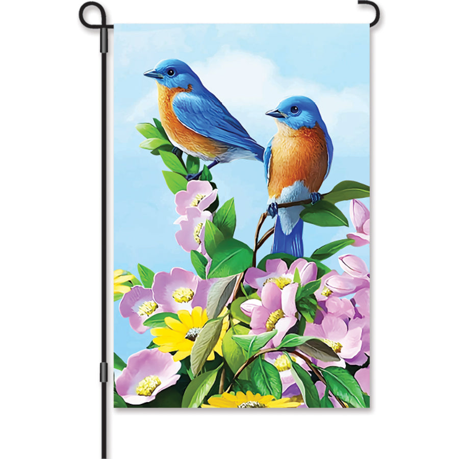 12 in. Floral Bluebirds garden flag featuring bright bluebird illustrations amidst vivid flowers, displayed outdoors on a flag stand.