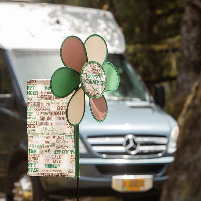Happy Camper Flower Spinner with colorful camping flag in a garden setting, featuring a vibrant flower design and pivoting pole.