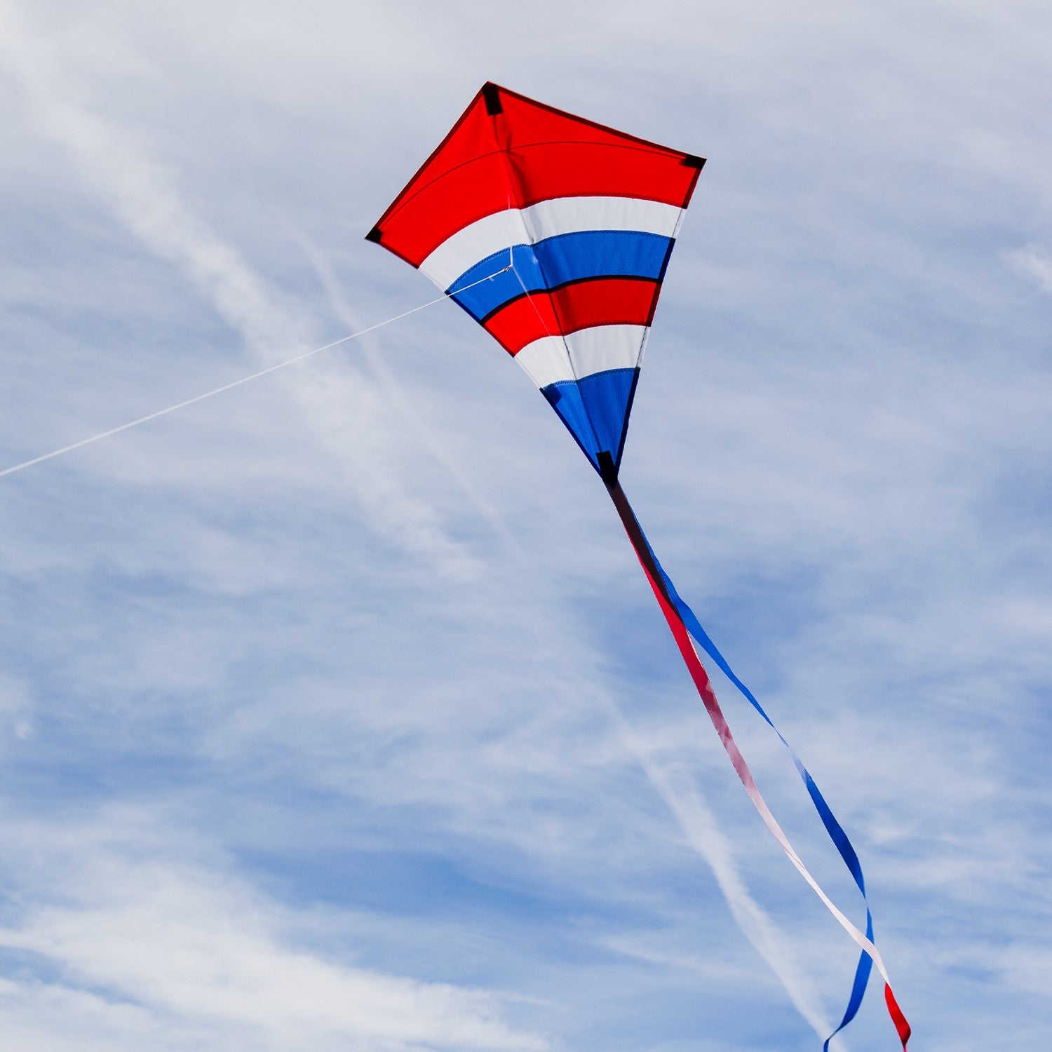 Alt Text: Patriotic Arch 27" Diamond Kite with vibrant colors and long tails flying against a blue sky during an outdoor family activity.
