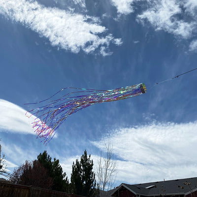Iridescent 51-inch Mylar windsock with holographic design fluttering in a gentle breeze, creating a colorful display in a garden setting.