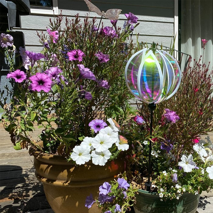 Iridescent 6-inch Gazing Ball Spinner with opalescent finish, reflecting sunlight in an outdoor garden setting, mounted on a sturdy fiberglass pole.