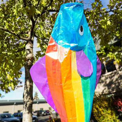 48-Inch Iridescent Rainbow Fishy Windsock featuring shimmering rainbow colors, purple fins, and a blue head, hanging in an outdoor garden.