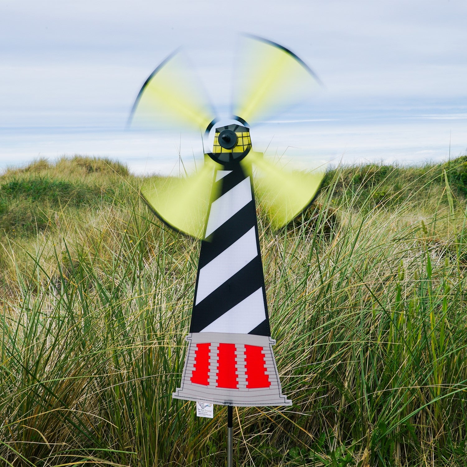 Lighthouse Spinner featuring Hatteras Lighthouse design, displayed outdoors with a colorful, rotating four-panel wheel in a garden setting.