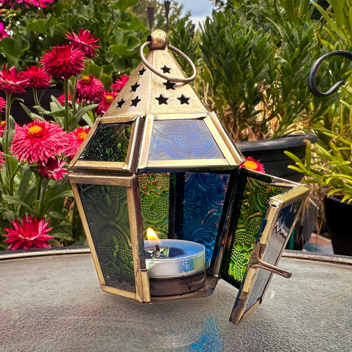 Mini Blue & Green Hex Glass Tealight Lantern with textured glass and brass accents hanging in an outdoor setting, radiating warm candlelight.