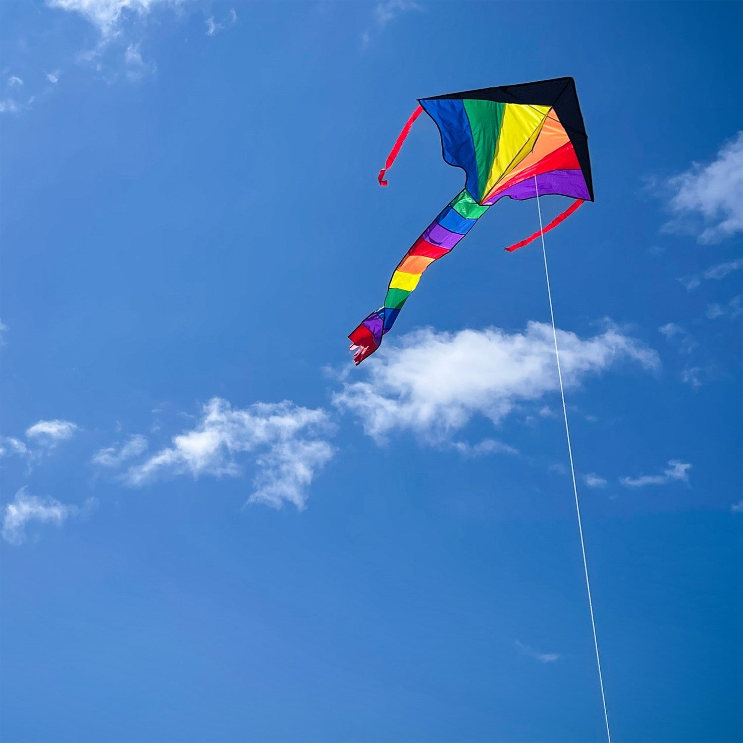 Brightly colored Mini Rainbow Fly-Hi Kite featuring a rainbow design flying in blue sky with streamer tails trailing behind it.