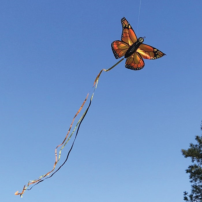 Monarch Butterfly Kite in vibrant orange and red colors soaring against a blue sky with tails fluttering gracefully behind it.