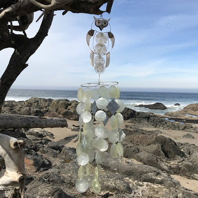 Owl Capiz Breeze Wind Chime made of handcrafted Capiz shells and textured metal displayed on a garden hook in a serene outdoor setting.