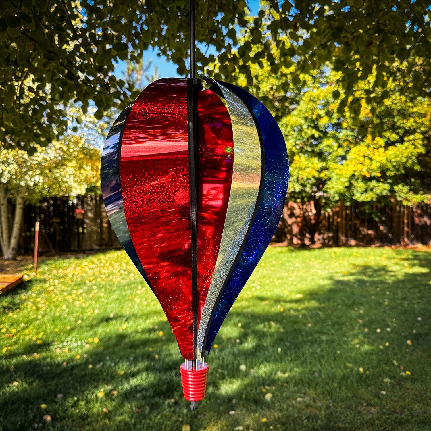 Patriot Sparkler 6-Panel Hot Air Balloon spinning in a garden, showcasing its vibrant red, white, and blue Mylar design against a blue sky.