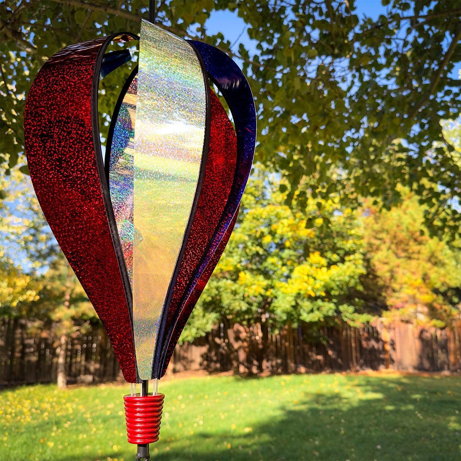 Patriot Sparkler 6-Panel Hot Air Balloon spinning in a garden, showcasing its vibrant red, white, and blue Mylar design against a blue sky.