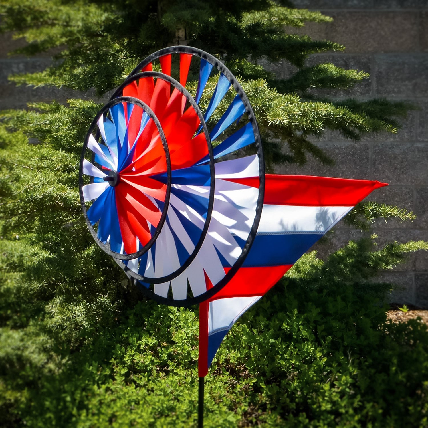 Patriot Wind Sail Triple Wheel Spinner with three colorful wheels and an applique windsail, displayed in an outdoor garden setting, showcasing red, white, and blue colors.