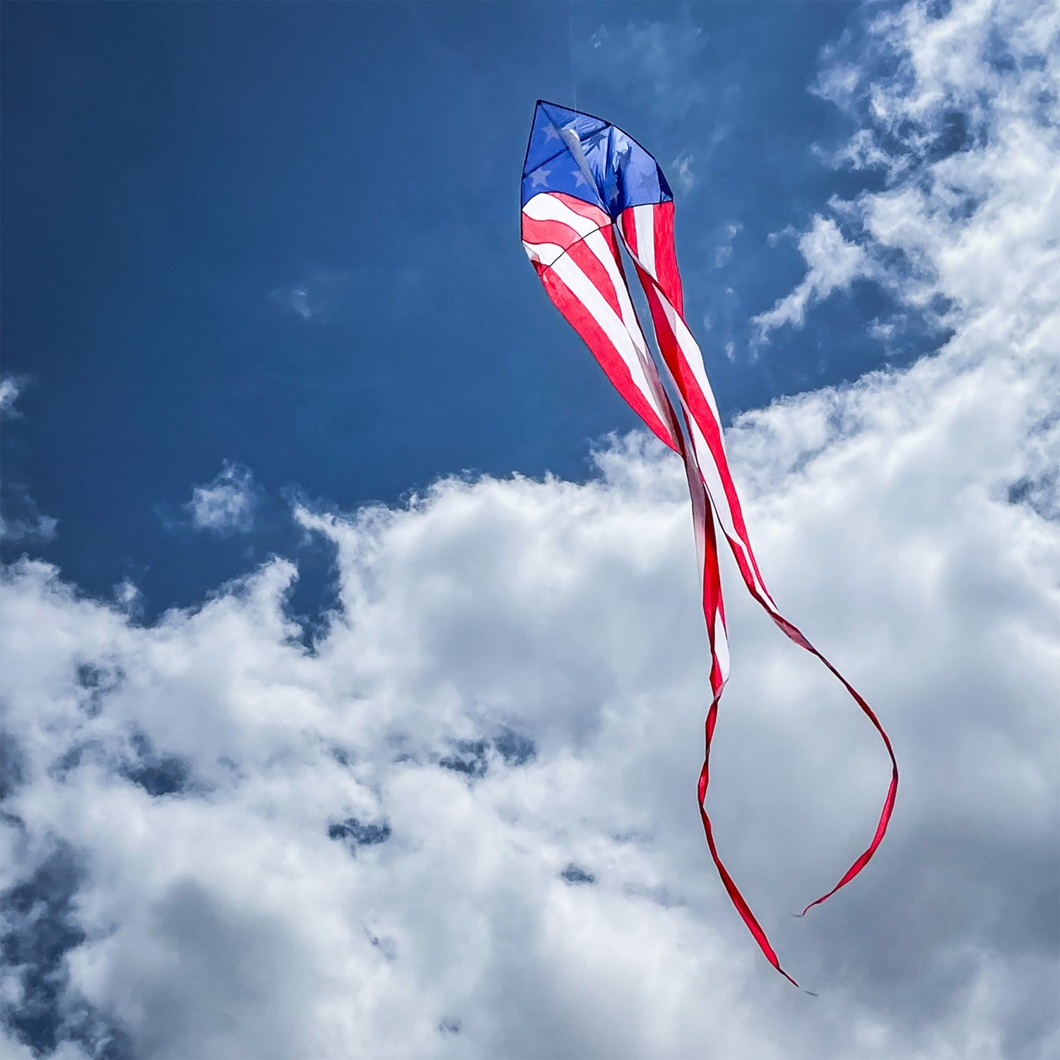 Patriotic 56" Delta kite featuring red, white, and blue stars and stripes with long tails, soaring in bright blue sky against a backdrop of fluffy clouds.