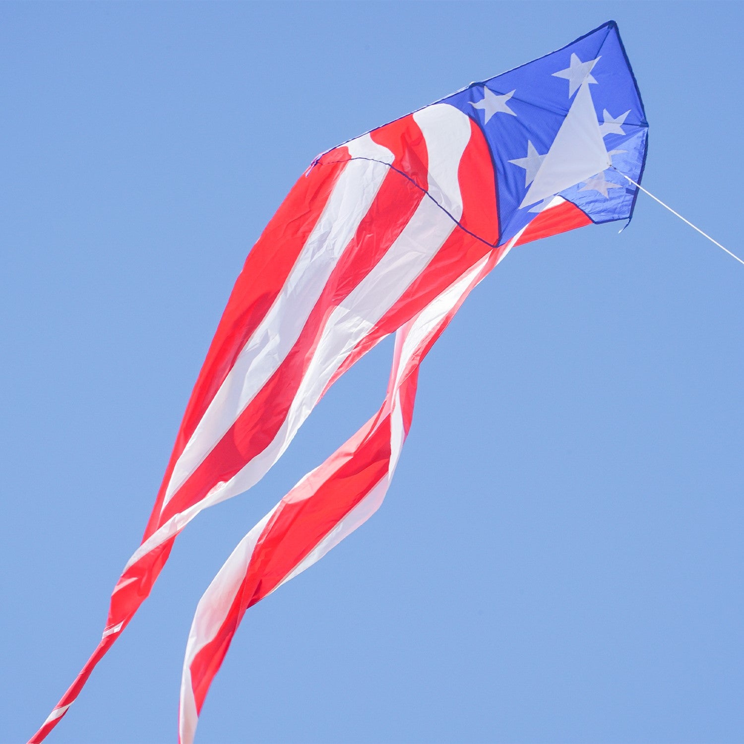 Patriotic 56" Delta kite featuring red, white, and blue stars and stripes with long tails, soaring in bright blue sky against a backdrop of fluffy clouds.