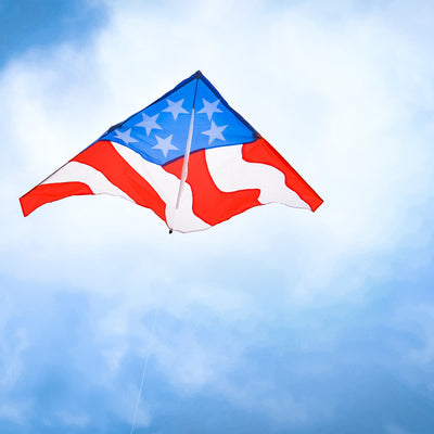 Patriotic 57 inch Delta Kite with red, white, and blue stars and stripes design soaring high in the blue sky against a sunny backdrop.