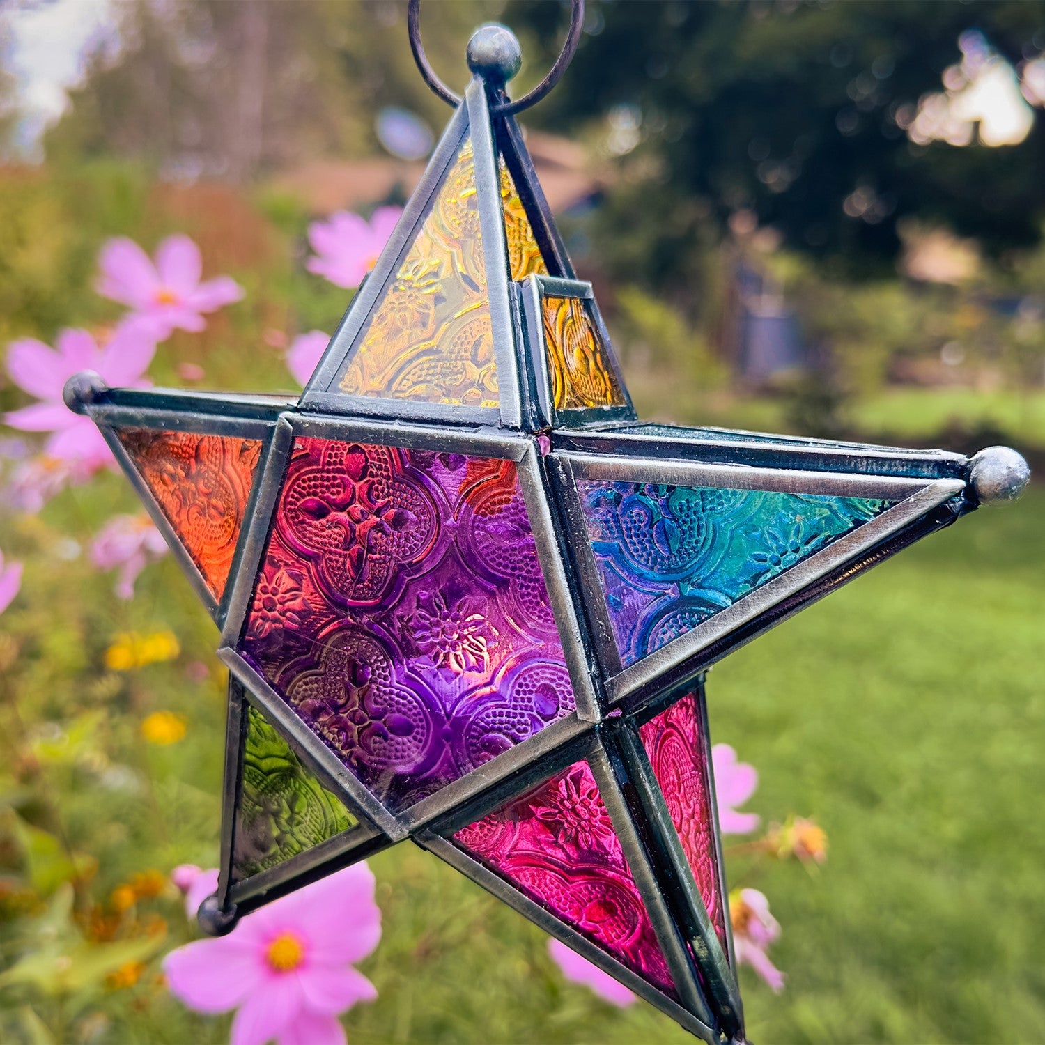 Rainbow 5-Point Star Hanging Tealight Lantern with multicolored glass panels and metal frame, displayed outdoors, radiating warm light from a tealight candle.