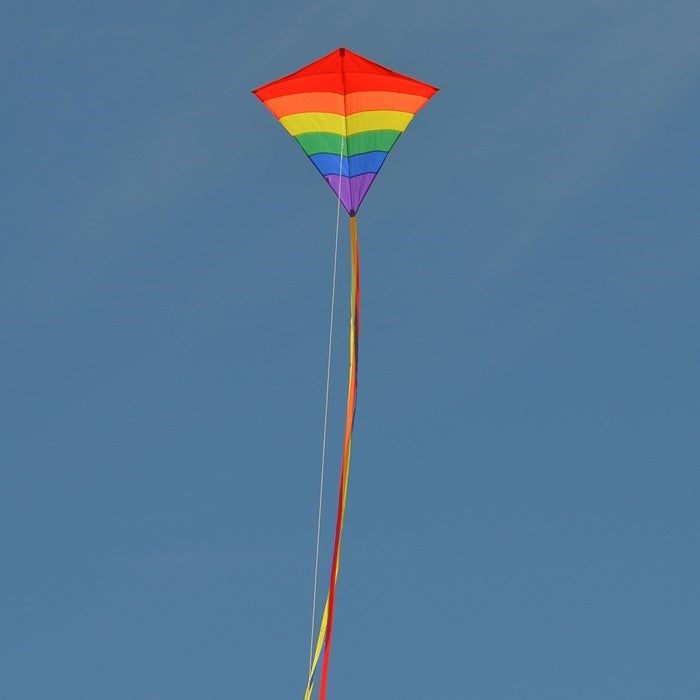 Rainbow Arch 39-Inch Diamond Kite in vibrant colors, featuring long fabric tails, soaring against a bright blue sky on a sunny day.