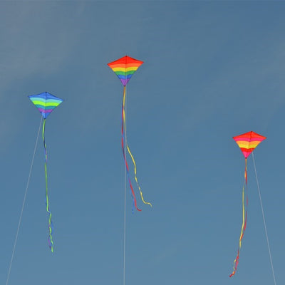 Rainbow Arch 39-Inch Diamond Kite in vibrant colors, featuring long fabric tails, soaring against a bright blue sky on a sunny day.