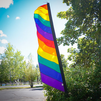 7-foot Rainbow Feather Banner with colorful applique stripes standing on an outdoor pole, illuminated by sunlight for a vibrant display.