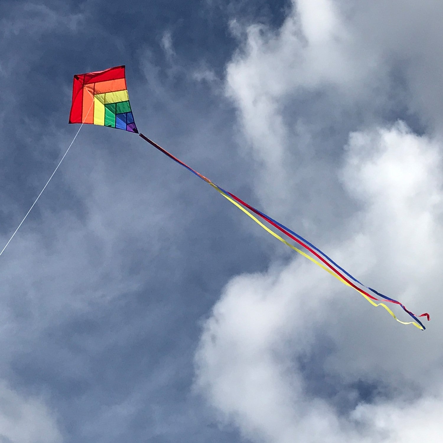 Rainbow Overlay 30-inch Diamond Kite flying in the blue sky with colorful streamer tails trailing below, showcasing its vibrant design.