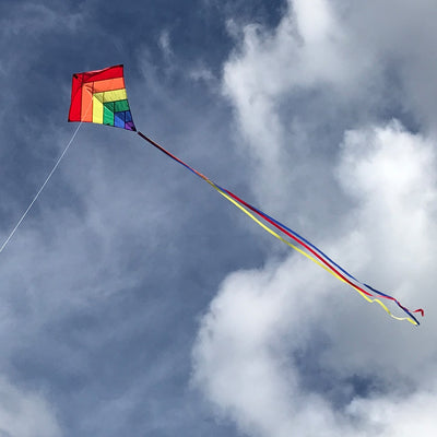 Rainbow Overlay 30-inch Diamond Kite flying in the blue sky with colorful streamer tails trailing below, showcasing its vibrant design.