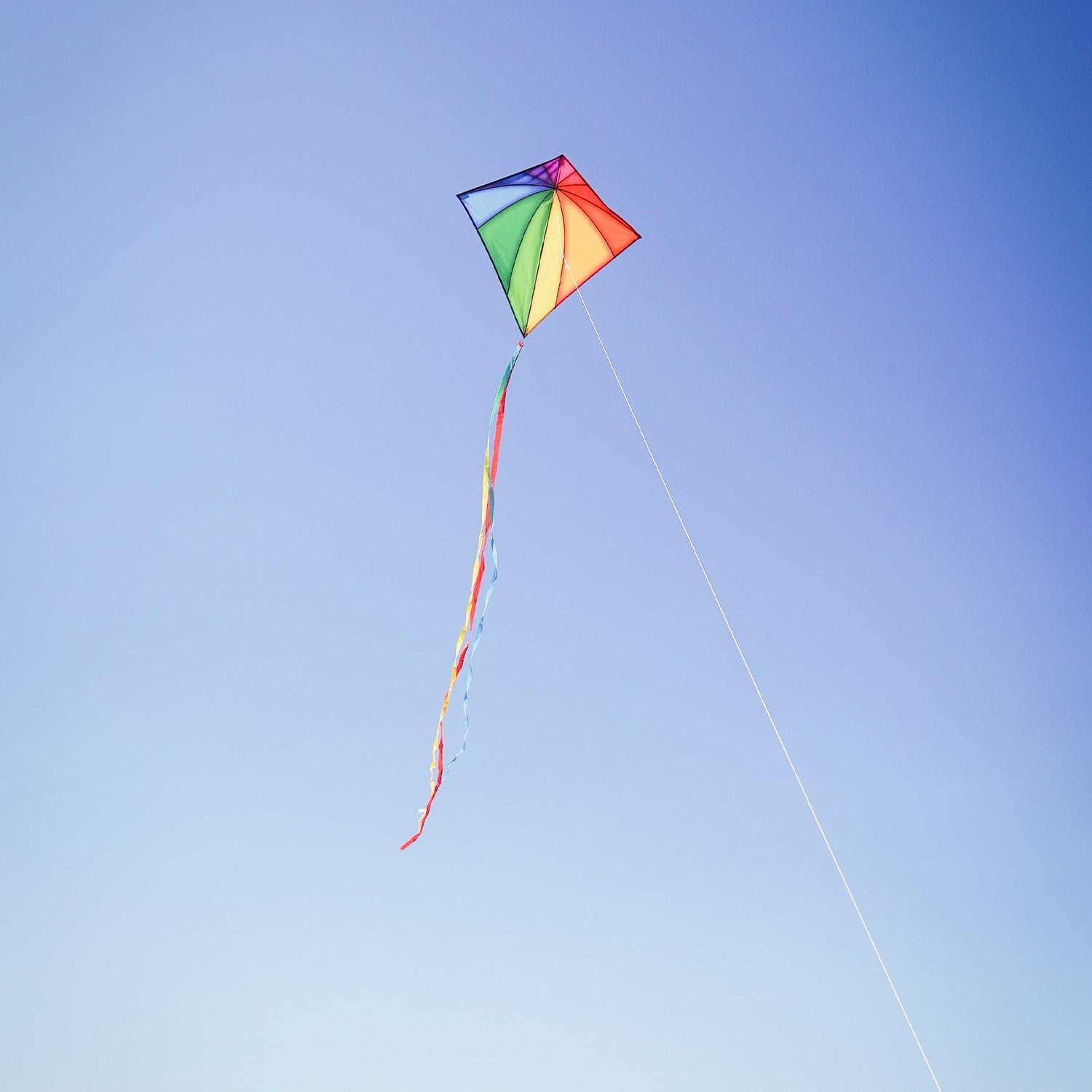 Colorful Rainbow Stained Glass 30-inch Diamond Kite flying against a blue sky, showcasing its vibrant stained glass patterns and attached tails.