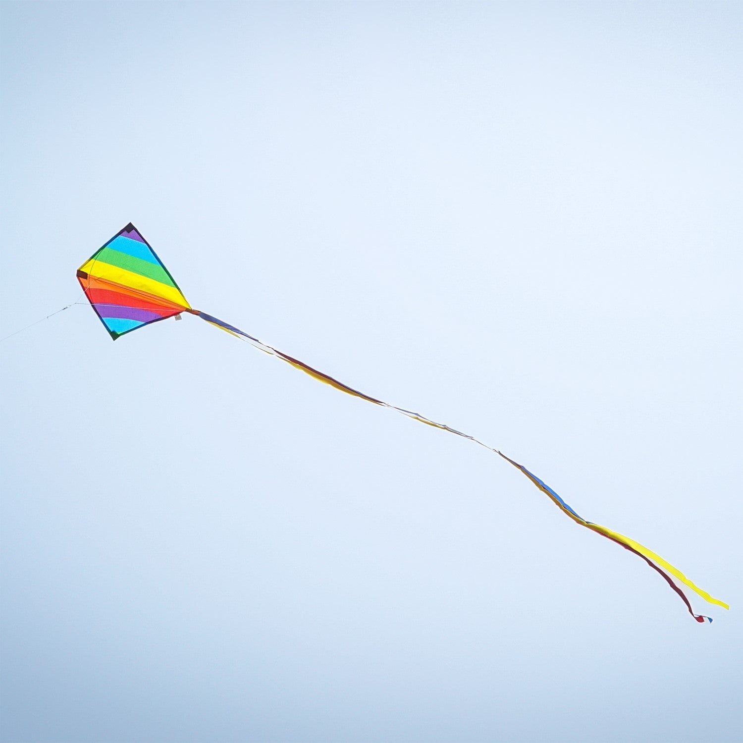 Rainbow Stripe 18" Diamond Kite flying high against a blue sky with colorful tails trailing behind, showcasing its vibrant spectrum design and classic shape.