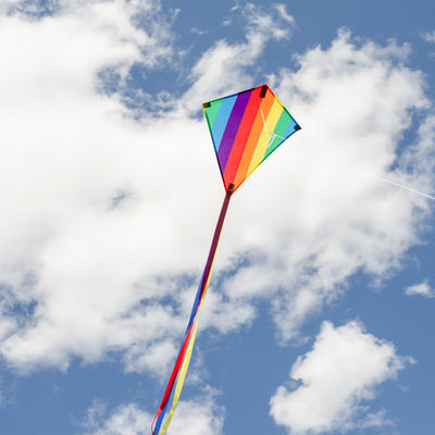 Rainbow Stripe 18" Diamond Kite flying high against a blue sky with colorful tails trailing behind, showcasing its vibrant spectrum design and classic shape.