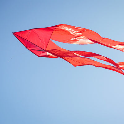Red 77' Wave Delta Kite soaring in the sky with long flowing tails visible, showcasing its vibrant color and impressive size against a clear backdrop.