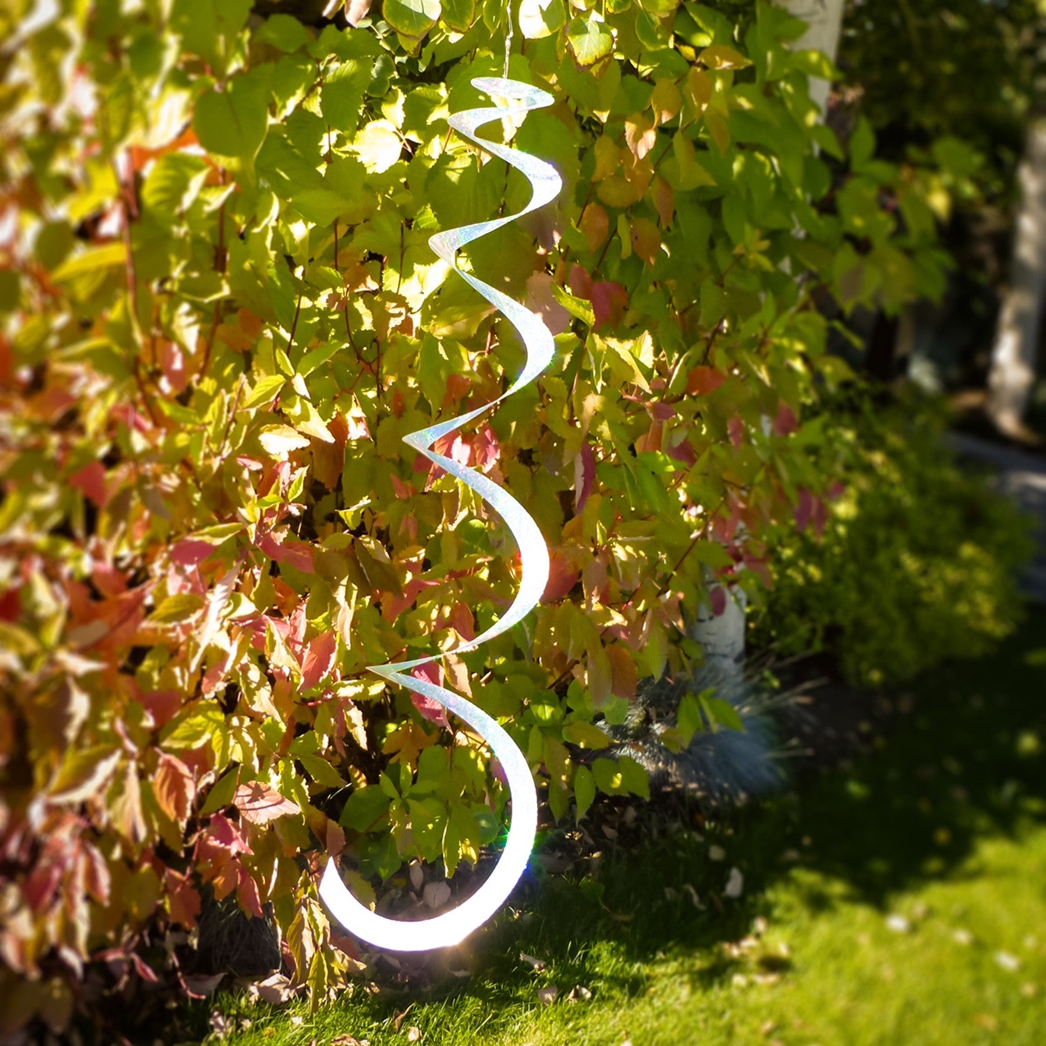 A Silver Sparkle Sprite hanging decor made from shimmering Mylar, gently twirling in a light breeze against a garden backdrop.