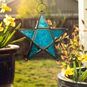 Teal 5-Point Star Hanging Tealight Lantern with textured glass panels and metal accents, suspended outdoors against a scenic backdrop.